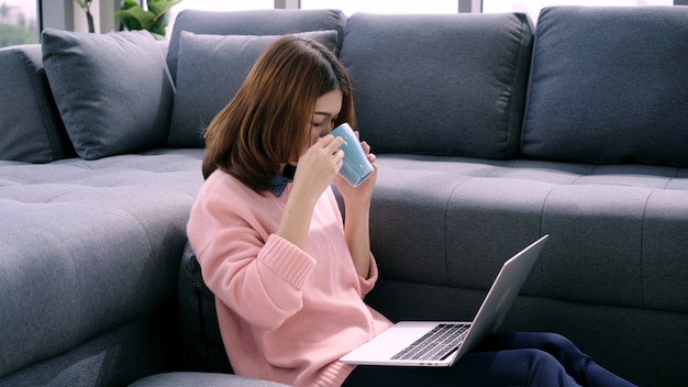 Портрет красивой привлекательной азиатской женщины используя компьютер или компьтер-книжку держа теплую чашку кофе или чая