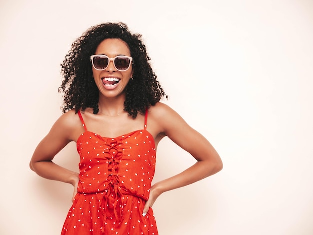 Portrait of beautiful black woman with afro curls hairstyle Smiling model dressed in red summer dress Sexy carefree female isolated white wall in studio