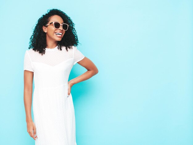 Ritratto di bella donna di colore con l'acconciatura di riccioli afro modello sorridente vestito in abito estivo bianco femmina spensierata sexy in posa vicino al muro blu in studio abbronzato e allegro