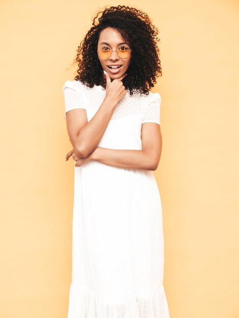 Portrait of beautiful black woman with afro curls hairstyle Smiling model dressed in white summer dress Sexy carefree female posing near yellow wall in studio Tanned and cheerful Isolated
