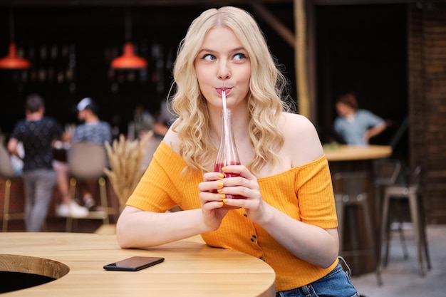 Free photo portrait of beautiful blond girl dreamily drinking lemonade in courtyard of city cafe