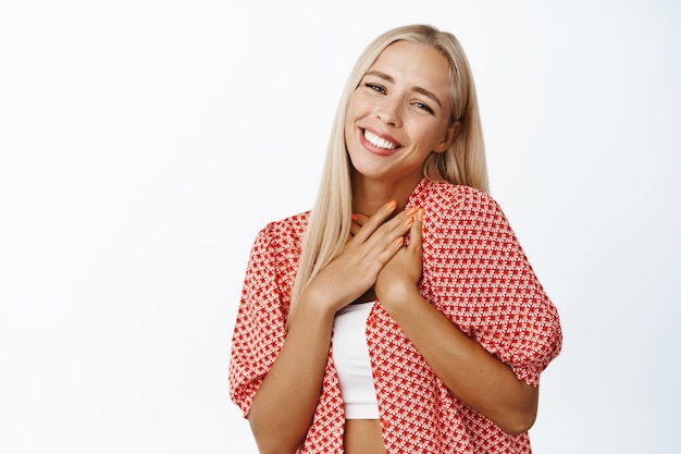 Free photo portrait of beautiful blond girl looking flattered and heartfelt holding hands on heart feeling romantic standing over white background