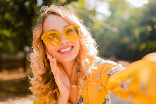 自撮り写真を作るサングラスを身に着けている黄色いブラウスで美しい金髪のスタイリッシュな笑顔の女性の肖像画