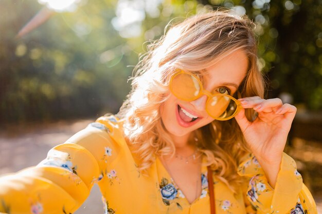 Portrait of beautiful blond stylish smiling woman in yellow blouse wearing sunglasses making selfie photo