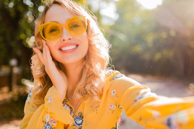 Portrait of beautiful blond stylish smiling woman in yellow blouse wearing sunglasses making selfie photo