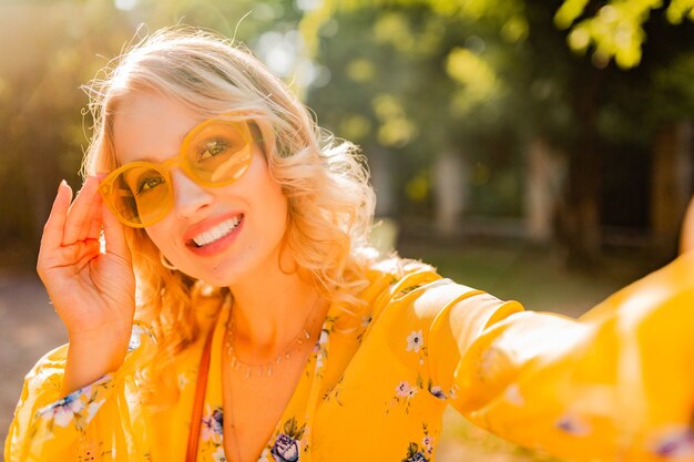 Portrait of beautiful blond stylish smiling woman in yellow blouse wearing sunglasses making selfie photo