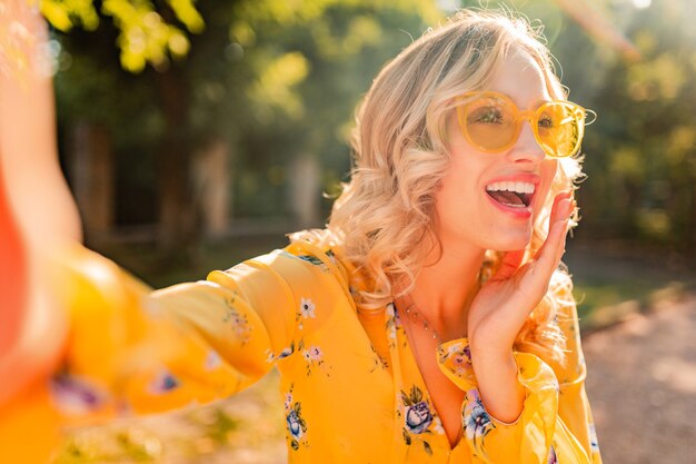 Portrait of beautiful blond stylish  smiling woman in yellow blouse wearing sunglasses making selfie photo