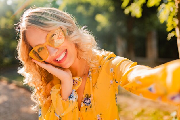 Portrait of beautiful blond stylish smiling woman in yellow blouse wearing sunglasses making selfie photo