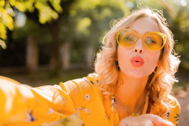 Portrait of beautiful blond stylish woman in yellow blouse wearing sunglasses taking selfie photo