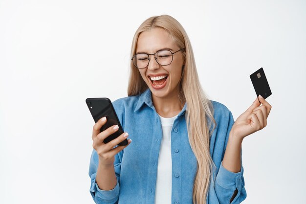 Portrait of beautiful blond woman in glasses laughing looking at mobile phone screen holding credit card shopping online standing over white background