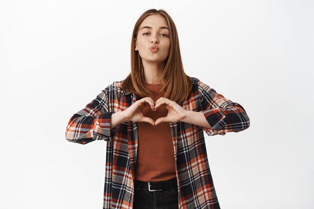 Portrait of beautiful blond woman kissing, show heart love sign, like someone, romantic date mood, sympathy towards someone, standing passionate and tender against white wall