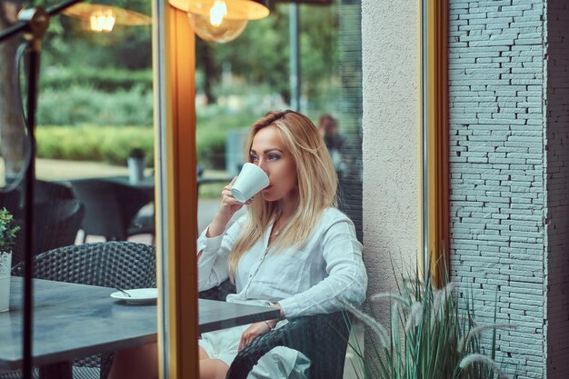 Portrait of a beautiful blonde female dressed in a white blouse sitting at a table drinks coffee at terrace cafe.