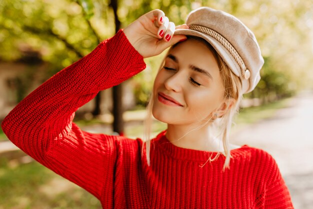 秋の公園でポーズをとって素敵な赤いセーターと軽い帽子の美しいブロンドの女の子の肖像画。