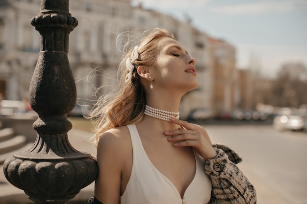 Free photo portrait of beautiful blonde lady in stylish white silk dress, checkered jacket and pearl necklace gently touching neck and posing at city square