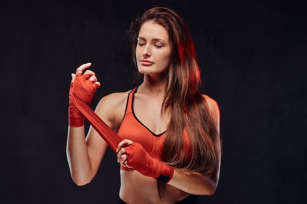 Portrait of a beautiful brunette female boxer in sports bra bandages her hands. Isolated on dark textured background.