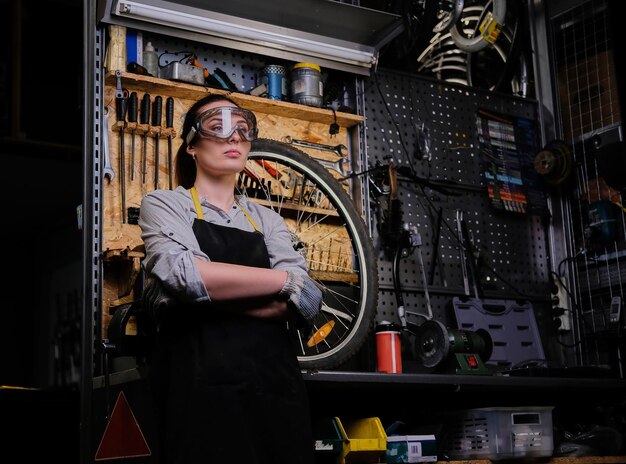 Portrait of a beautiful brunette female wearing working clothes, apron and goggles, standing with crossed arms in a workshop against wall tools.