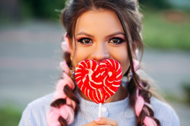 Portrait of beautiful brunette girl looking at camera, hiding face by candy red heart on stick. Pretty and stylish woman with make up and colorful pink braids. Romantic playful mood.
