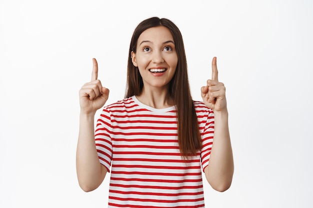 Portrait of beautiful brunette girl pointing, looking up and smiling happy, reading promotional text, showing sale banner, standing in red t-shirt against white background.