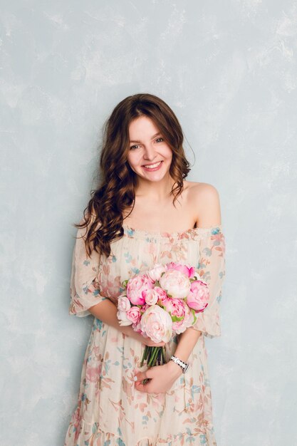 Portrait of a beautiful brunette girl with curly hair standing in a studio. She wears light silk dress with open shoulder and holds flowers in her hands. She has white watch on her wrist. She smiles.