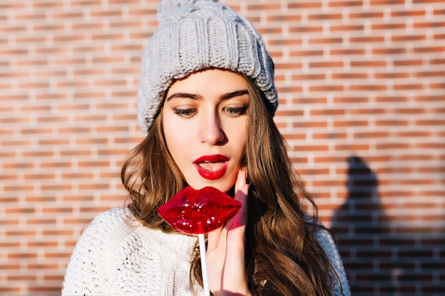 Portrait beautiful brunette girl with long hair in knitted hat on wall  outside. She looks astonished at lollipop red lips.