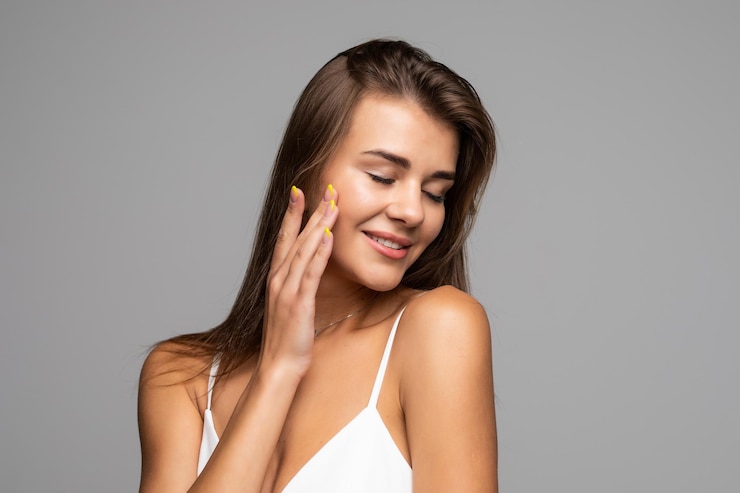 Free Photo portrait of beautiful brunette with bare shoulders touching her cheek, looking at camera and smiling on gray