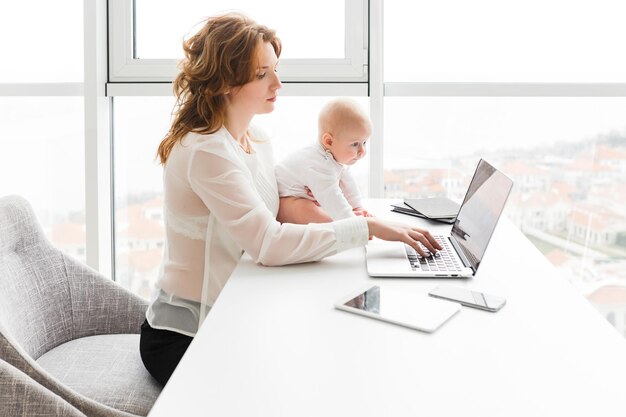 Portrait of beautiful business woman holding her cute little baby while sitting at the table and working on laptop