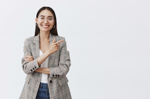 Portrait of beautiful carefree adult woman in glasses and jacket over white t-shirt, pointing at upper right corner and smiling with shiny teeth