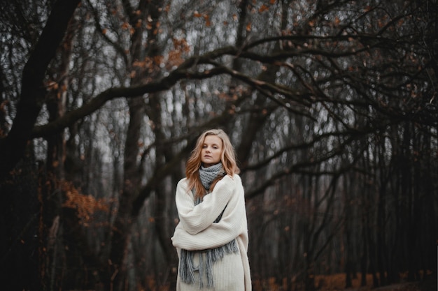 Ritratto di bella ragazza bionda caucasica vestita di pullover bianco, arricciata da sola nella foresta autunnale