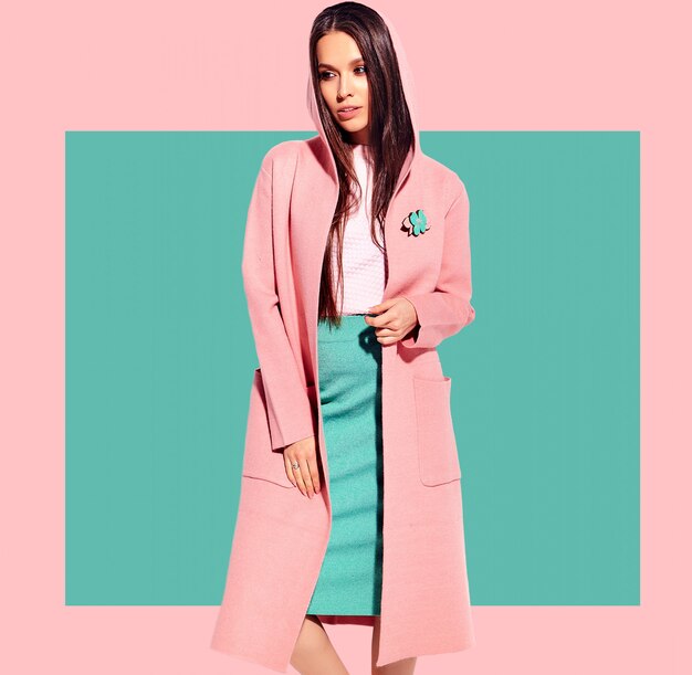 Portrait of beautiful caucasian smiling brunette woman model in bright overcoat and summer stylish skirt posing  on pink and blue background