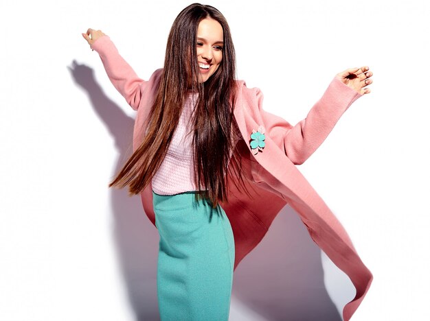 Портрет красивой кавказской улыбающейся брюнетки модели в ярко-розовом пальто и летней стильной синей юбке на белом фоне
