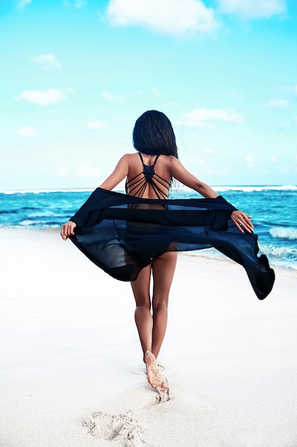 青い空と海に白い砂と夏のビーチでポーズをとって飛行岬と黒の水着で黒い長い髪の美しい白人日光浴女性モデルの肖像