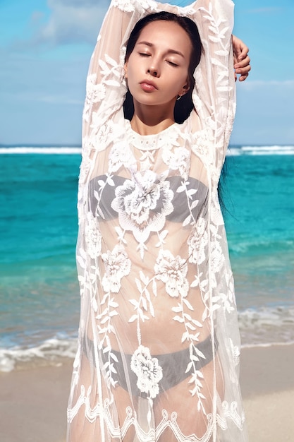 Free photo portrait of beautiful caucasian woman model with dark long hair in transparent white long blouse dress posing on summer beach with white sand on blue sky and ocean