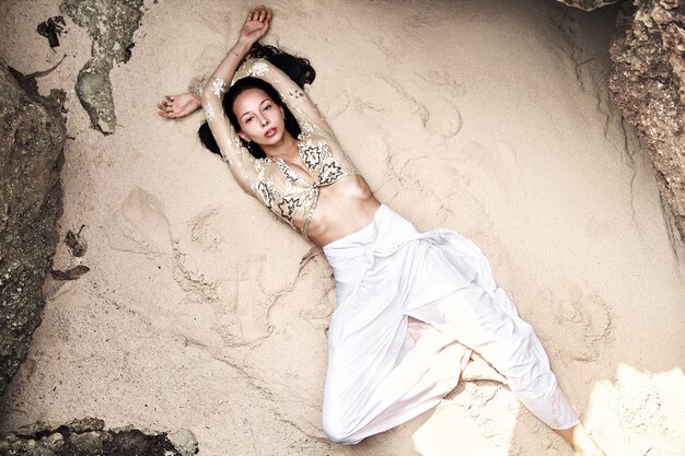 portrait of beautiful caucasian woman model with dark long hair in wide-leg classic pants  lying on white sand on the beach near rocks. Top view
