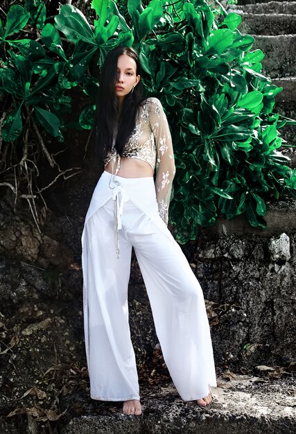 portrait of beautiful caucasian woman model with dark long hair in wide-leg classic pants  posing near green tropical exotic leaves background