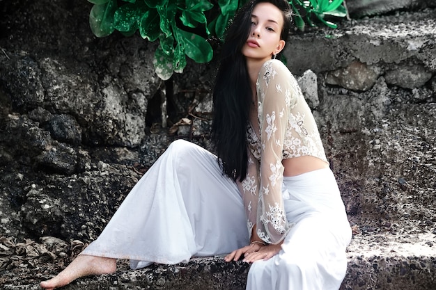 portrait of beautiful caucasian woman model with dark long hair in wide-leg classic pants  posing near rocks and green tropical exotic leaves background
