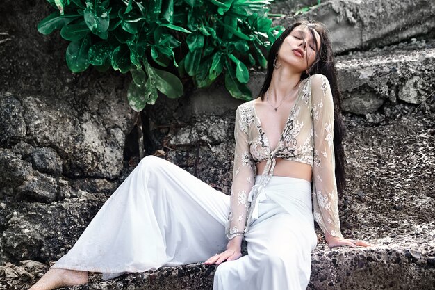 portrait of beautiful caucasian woman model with dark long hair in wide-leg classic pants  posing near rocks and green tropical exotic leaves background
