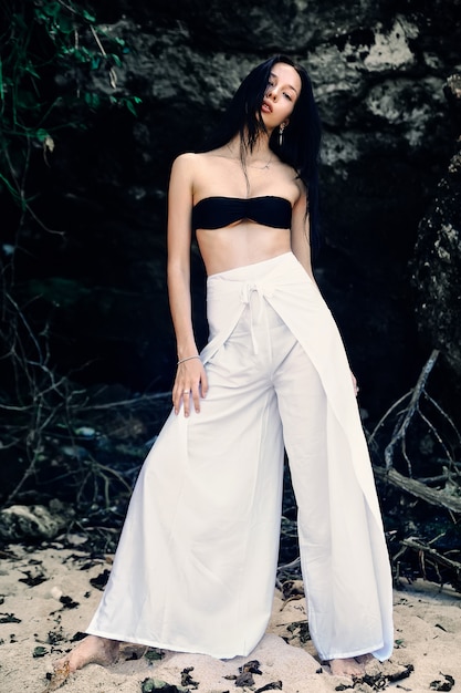 Free photo portrait of beautiful caucasian woman model with dark long hair in wide-leg classic pants posing near rocks on summer beach