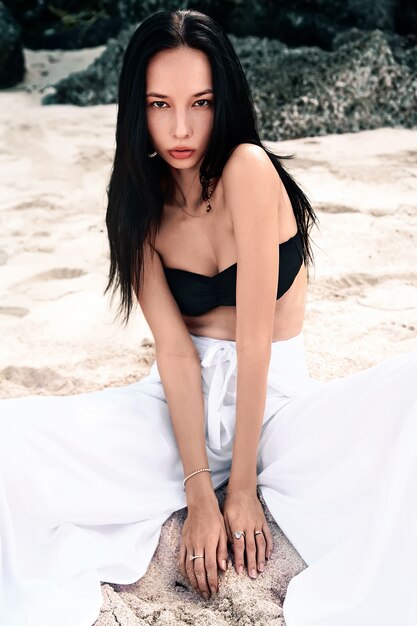 portrait of beautiful caucasian woman model with dark long hair in wide-leg classic pants sitting on summer beach with white sand near rocks