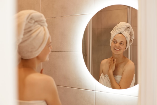 Foto gratuita ritratto di bella donna caucasica con un asciugamano sui capelli che si tocca il collo in bagno, guardando il suo riflesso nello specchio, in posa dopo aver fatto la doccia.