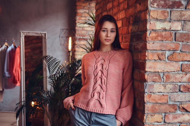 Foto gratuita ritratto di una bella bruna affascinante vestita con abiti eleganti sorridente mentre si appoggia a un muro di mattoni in una stanza con interni soppalcati.