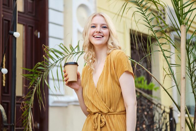 Portrait of beautiful cheerful blond girl in dress with coffee to go joyfully looking in camera on city street