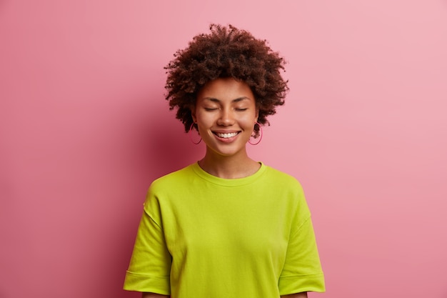 Portrait of beautiful cheerful woman closes eyes and smiles with pleasure, wears casual green t shirt, hears pleasant words of support, isolated in pink wall. Happy emotions and feelings