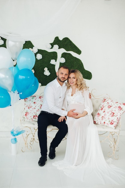 Portrait of beautiful couple of mom and dad embracing stomach and looking each other touching face to face Pregnant blonde woman in white smiling to her handsome husband posing at stylish interior