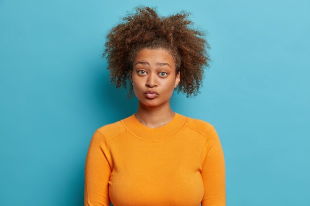 Portrait of beautiful curly haired Afro American woman keeps lips rounded makes funny grimace dressed in casual orange jumper.