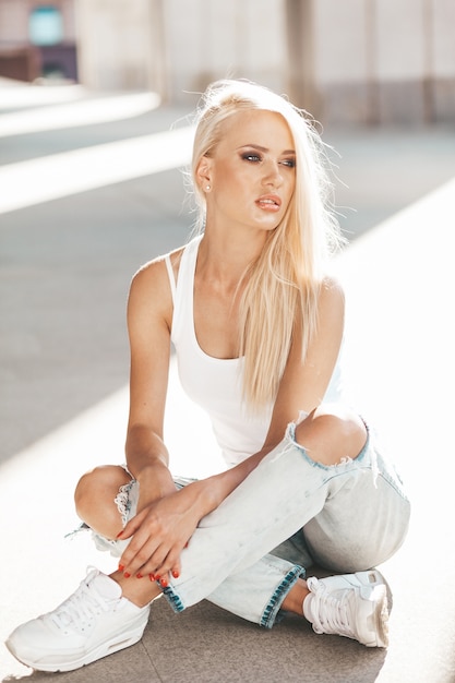 Портрет красивой милой белокурой девушки в белой футболке и джинсах представляя outdoors. Милая девушка сидит на асфальте на улице