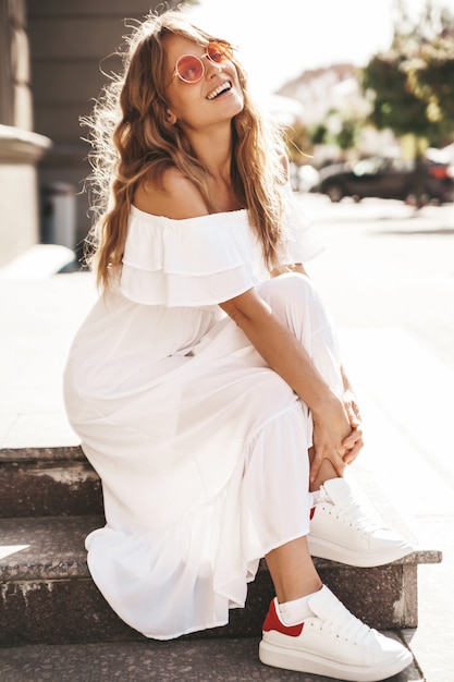 Портрет красивой милой белокурой модели подростка без макияжа в летней хипстерской белой одежде, сидящей на фоне улицы