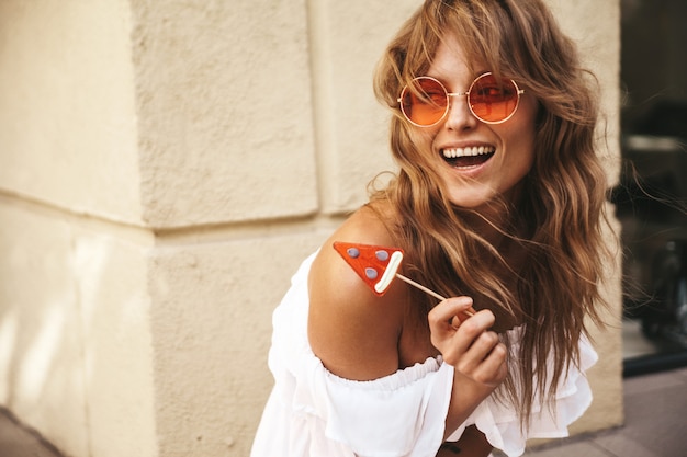 Free photo portrait of beautiful cute blond teenager model with no makeup in summer hipster white dress clothes with watermelon candy posing near yellow wall