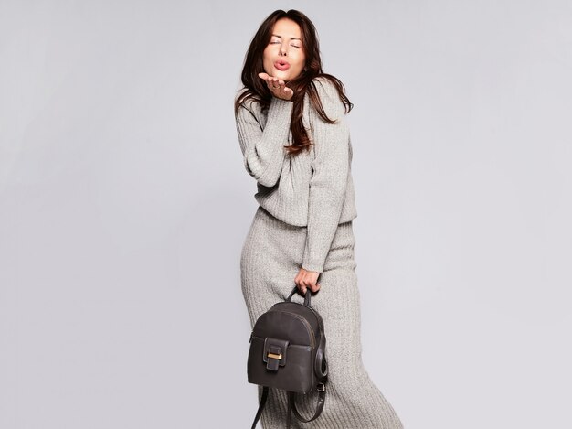 핸드백와 회색에 고립 된 화장없이 캐주얼가 회색 스웨터 옷에 아름 다운 귀여운 갈색 머리 여자 모델의 초상화. 키스하기