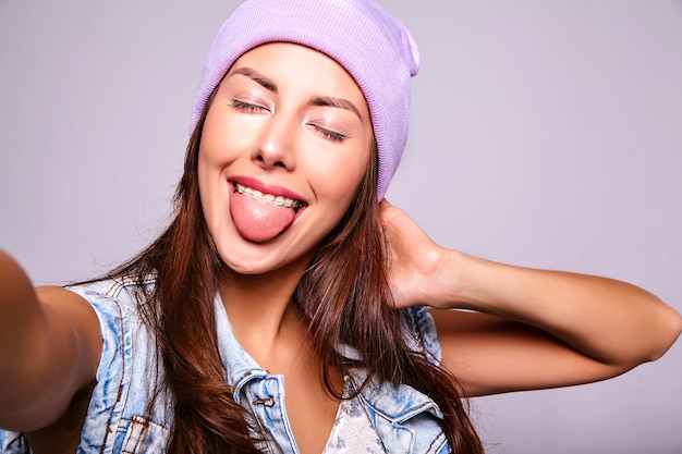 Free photo portrait of beautiful cute brunette woman model in casual summer jeans clothes with no makeup in purple beanie making selfie photo on phone isolated on gray. showing her tongue