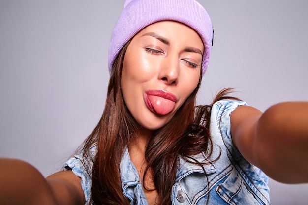 Il ritratto di bello modello sveglio della donna castana in jeans casuali dell'estate copre senza trucco in berretto porpora che fa la foto del selfie sul telefono isolato su gray. mostrando la sua lingua